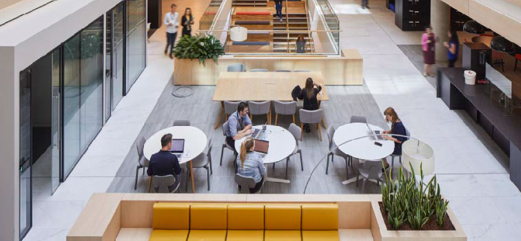 A common area inside the main Central Bank of Ireland offices with a yellow sofa and three round tables with four people seated and looking at laptops are in the foreground. A table, five people and stairs are the background area.